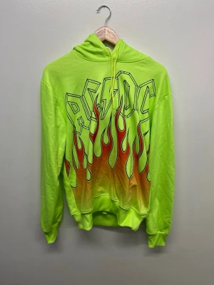 Neongrön AC/DC hoodie med flames - Cool neongrön hoodie med stor AC/DC-logga och eldflammor i rött och orange på framsidan. Tröjan har huva med dragsko och ribbade muddar. Perfekt för dig som vill sticka ut och gillar rockig stil.
