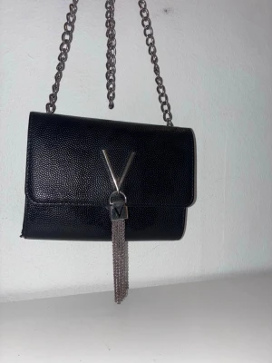 Svart axelväska med kedja från Valentino - Snygg svart axelväska från Valentino med silverfärgad kedjerem och ikoniskt V-emblem framtill. Väskan har en elegant rektangulär form och dekorativa silverfransar som hänger från låset. Perfekt för dig som gillar stilrena detaljer.
