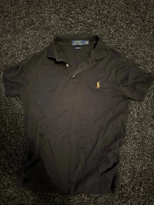 Svart pikétröja från Polo Ralph Lauren - Klassisk svart pikétröja från Polo Ralph Lauren den har lite stretchigare material en de vanliga pikétröjorna så perfekt för dig som letar efter en tröja med perfekt passform