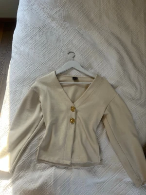 Beige kofta med guldknappar - Stilren beige kofta från Gina med två stora guldknappar framtill. Koftan har v-ringning, lång ärm och figurnära passform. Perfekt för att lyfta en enkel outfit med en touch av elegans.