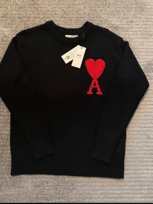 Svart stickad tröja från Ami Paris - Svart stickad tröja från Ami Paris med röd hjärtlogga och bokstaven A på bröstet. Tröjan har rund halsringning, ribbade muddar och är tillverkad i mjukt material. Perfekt för en trendig och avslappnad look.