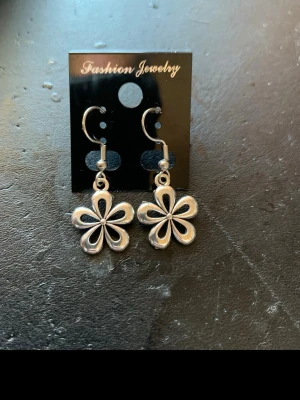 Silvriga blommiga örhängen - Säljer ett par silvriga örhängen i form av blommor med fem kronblad. De har en blank finish och hängande krokfäste. Perfekta för dig som vill ha en söt och stilren accessoar till din look.