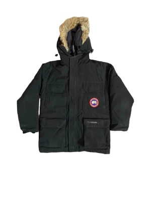 Svart parkas från Canada Goose - Svart parkasjacka från Canada Goose med huva och beige fuskpäls. Jackan har flera stora fickor framtill, klassisk logotyp på bröstet och justerbar huva. Perfekt för kalla dagar och har en robust design som håller värmen.