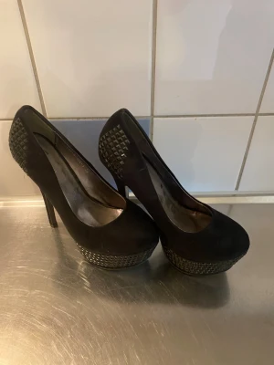 Svarta pumps med nitar från Nelly - Snygga svarta pumps från Nelly med hög klack och platåsula. Skorna har coola nitar på hälen och runt sulan som ger en edgy look. Tillverkade i ett mockaliknande material och har rundad tå. Perfekta för dig som vill sticka ut lite extra.