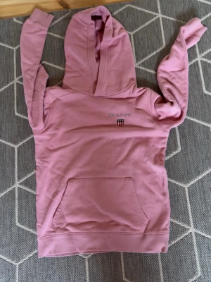Rosa hoodie från GANT med huva - Säljer en rosa hoodie från GANT med broderad logotyp och sköld på bröstet. Hoodien har en stor huva och en praktisk magficka framtill. Materialet är mjukt och bekvämt, perfekt för en avslappnad stil. strl 11-12 men passar 156-168