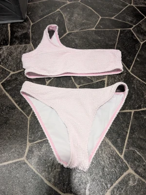 Ljusrosa bikini med textur stl 160 - Trendig ljusrosa bikini med en axelbandstopp och matchande bikinitrosa. Materialet har en ribbad, frottéliknande textur som ger en unik look. Perfekt för sommarens stranddagar eller poolhäng.