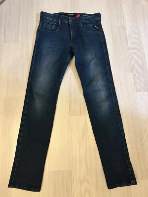 Mörkblå slim fit jeans från Replay 30/31 - Snygga mörkblå slim fit jeans från Replay med klassisk femficksdesign och diskreta slitningar framtill. Jeansen har raka ben och tydliga sömmar, perfekt för en stilren och modern look.