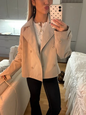Beige kort trenchcoat  - Säljer en stilren beige kort trenchcoat med bred krage och dubbelknäppning. Jackan har en klassisk design med stora knappar och raka ärmar. Perfekt för dig som vill ha en enkel och snygg look.