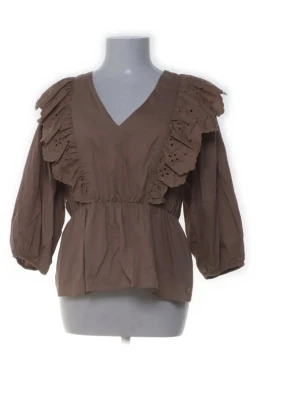Brun volangblus med v-ringning - Snygg brun blus med breda volanger och broderade detaljer längs axlarna. Blusen har v-ringning, trekvartsärm och resår i midjan för en feminin siluett. 