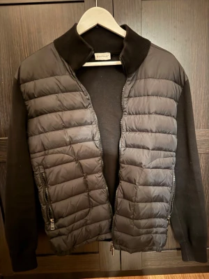 Svart dunväst från Moncler - Stilren svart dunväst från Moncler med quiltad front, ribbad krage och ärmar i mjukt material. Två dragkedjeförsedda fickor och dubbel dragkedja framtill. Perfekt för lager-på-lager och en clean look. NFC fungerar. Pris kan diskuteras. Jackan passar inte mig mer.