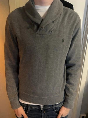Grå tröja med sjalkrage från Polo Ralph Lauren - Snygg grå tröja från Polo Ralph Lauren med sjalkrage. Strlk M. Schyssta knappar på kragen. Gott skick. Pris går att diskutera