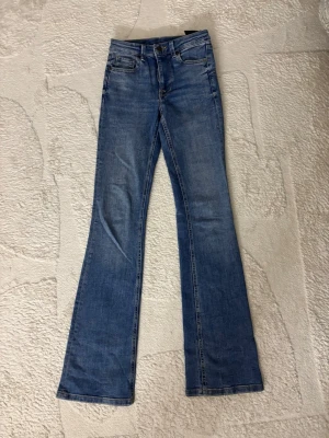 Blå bootcut jeans med utsvängda ben - Säljer ett par klassiska blå bootcut jeans med utsvängda ben och fem fickor. Jeansen har normal midja och är gjorda i ett mjukt denimtyg. Perfekta för dig som gillar retrovibbar och vill ha en avslappnad men snygg look.