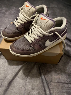 Nike SB Dunk Low lila/vit sneakers - Nike SB Dunk Low sneakers i lila och vitt med vit swoosh och orange Nike-logga på plösen. Ovandelen är i en mix av mocka och textil, med vita skosnören och mellansula. Klassisk låg siluett som passar till streetwear och skate.
