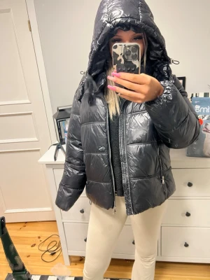 Svart pufferjacka från 157 med huva - Säljer en svart pufferjacka från 157 med glansigt material och quiltad design. Jackan har en stor huva, dragkedja framtill och är perfekt för kalla vinterdagar. Enkel och stilren look som passar till många outfits.