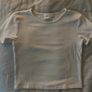 Vit kortärmad topp från Gina Tricot - Enkel och stilren vit topp från Gina Tricot i storlek 158/164. Modellen är kortärmad med rund halsringning och har en rak, något croppad passform. Perfekt att matcha med jeans eller kjol för en clean look.