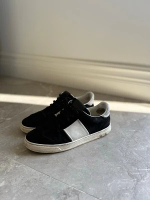 Valentino flycrews - Säljer ett par svarta sneakers med vit panel på sidan och vita detaljer vid häl och sula. Skorna har lågt skaft, svarta skosnören och en klassisk siluett. Materialet är en mix av mocka och läderimitation. Perfekta för dig som gillar stilrena och tidlösa sneakers. 