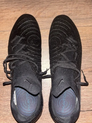 Svarta Nike fotbollsskor med mönster - Säljer ett par svarta Nike fotbollsskor med snyggt mönster på ovandelen och klassisk snörning. Skorna har en strumpliknande passform och är tillverkade i textilmaterial för bra komfort på planen. Perfekta för dig som vill ha stilrena och funktionella skor. Påsen kommer med.