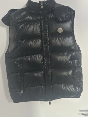 Svart dunväst från Moncler - Snygg svart dunväst från Moncler med glansig finish och ikonisk logga på bröstet. Västen har två dragkedjeförsedda fickor, hög krage och avtagbar huva. Perfekt för lager-på-lager och riktigt skön att bära under kyliga dagar.