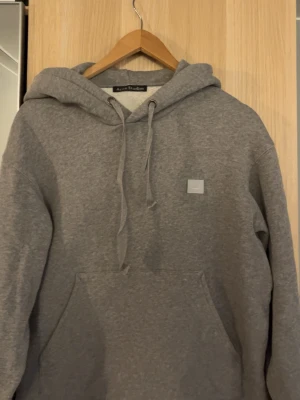 Grå hoodie från Acne Studios - Snygg grå hoodie från Acne Studios med klassisk huva och dragsnören. På bröstet sitter Acne Studios ikoniska face-patch. Hoodien har en stor magficka och är tillverkad i mjukt bomullsmaterial. Perfekt för en avslappnad och trendig stil. Äkta vara!
