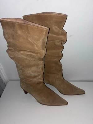 Beige slouchy stövletter i mocka Bukela - Säljer ett par snygga beige stövletter i mocka med slouchy skaft och spetsig tå från Bukela. Skorna har en låg, bred klack och en stilren design som ger en trendig vibe. Perfekta för dig som vill ha boots med lite extra attityd och mjukt material.