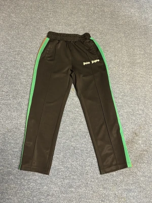 Palm Angels svarta mjukisbyxor - Svarta mjukisbyxor från Palm Angels med gröna sidoränder och resår i midjan. Byxorna har fickor och logga i vitt på vänster sida. Snygg streetwear-look med raka ben och sportig känsla. Perfekta för en avslappnad stil.