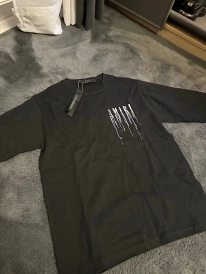 Svart AMIRI t-shirt med tryck - Svart t-shirt från AMIRI med färgstarkt tryck framtill och stort AMIRI-tryck med färgstreck på ryggen. Klassisk passform och rund halsringning. Perfekt för dig som gillar streetwear och vill sticka ut med en unik design.