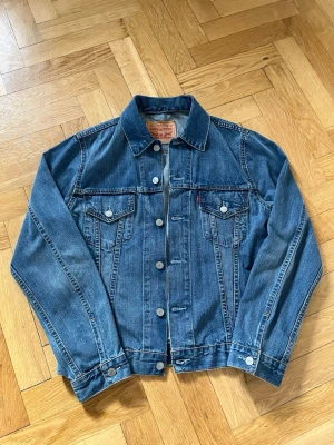 Klassisk blå jeansjacka från Levi's - Snygg blå jeansjacka från Levi's med raka linjer, knäppning framtill och två bröstfickor med lock. Jackan har klassisk krage och detaljerade sömmar. Perfekt för dig som gillar tidlös streetstyle. Storlek M men passar även S. Priset är diskuterbart.
