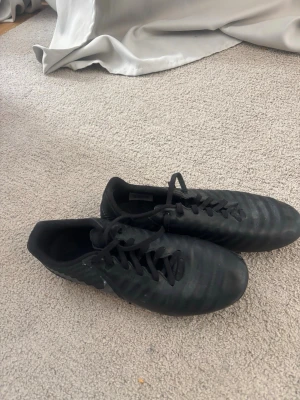 Svarta fotbollsskor från Nike - Säljer ett par helsvarta fotbollsskor från Nike med klassisk design och snörning. Skorna har dobbar för spel på gräs och är tillverkade i syntetmaterial med en stilren look. Perfekta för dig som vill ha en clean och sportig stil på planen. Storleken är 41, men dom passar även för dom som har storlek 38 och 39 