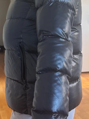 Svart pufferjacka med hög krage - Snygg svart pufferjacka med glansig finish och hög krage. Jackan har en quiltad design, dragkedjeficka på sidan och breda kanaler för extra värme. Perfekt för kalla dagar när du vill hålla stilen och värmen. Köpte den som storlek 16. Skulle säga att det motsvarar xs/s.