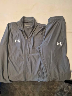 Grått träningsset från Under Armour - Säljer ett grått träningsset från Under Armour med dragkedja på jackan och resår i midjan på byxorna. Setet har vita loggor på både jacka och byxa och är gjort i ett mjukt, stretchigt material som passar perfekt för träning.