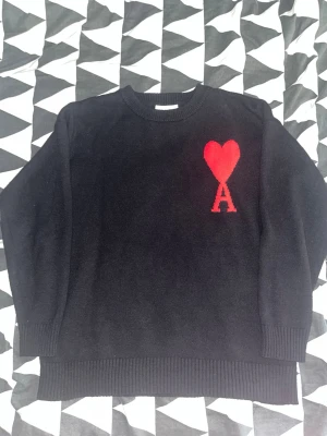 Svart stickad tröja Ami  - Säljer en svart stickad tröja från Ami med röd hjärtlogga och bokstaven A på bröstet. Tröjan har rund halsringning, ribbade muddar och är gjord i mjukt material. Perfekt för dig som gillar stilrena plagg med en twist. medium passar large