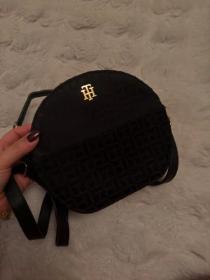 Svart handväska från Tommy Hilfiger - Snygg svart handväska från Tommy Hilfiger med guldfärgad logga framtill. Väskan har rundad form, justerbar axelrem och diskret TH-mönster i tyget. Perfekt för dig som gillar stilrena accessoarer med en lyxig touch.