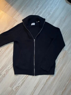 Svart stickad ziptröja Jack & Jones - Säljer en svart stickad ziptröja från Jack & Jones. Tröjan har hög krage, grovstickad struktur och dragkedja framtill. Perfekt för lager på lager och enkel att matcha till olika outfits.