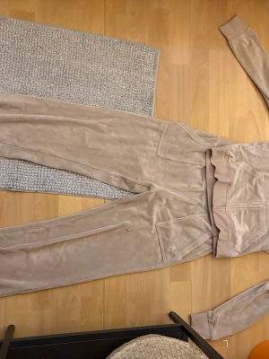 Beige velourset från Juicy Couture - Mysigt loungewear-set i beige velour från Juicy Couture. Setet består av en zip-hoodie och matchande byxor med resår i midjan och fickor. Perfekt för chill dagar hemma eller när du vill vara extra bekväm.