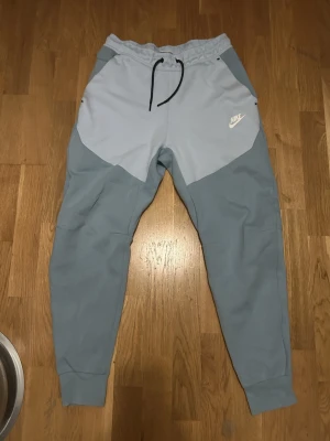 Nike ljusblå mjukisbyxor med snörning - Nike mjukisbyxor i två nyanser av ljusblått med svart snörning i midjan och vit Nike-logga på vänster sida. Byxorna har elastisk midja och muddar vid bensluten för en skön och avslappnad passform. Perfekta för chill eller träning.