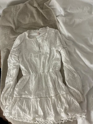 Vit spetsklänning från Nelly - Supersöt vit kortklänning från Nelly med broderade spetsdetaljer och volangkant. Klänningen har långärmade ärmar, markerad midja och v-ringad hals med dekorativ kant. Perfekt för sommarens alla tillfällen!