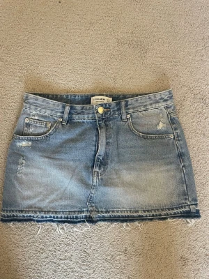 Blå jeanskjol med fransig kant - Snygg blå jeanskjol från Pull&Bear med klassisk femficksdesign, slitna detaljer och fransig nederkant. Kjolen har bälteshällor och knappstängning framtill. Perfekt för en avslappnad och trendig look. Använt fåtal gånger. 