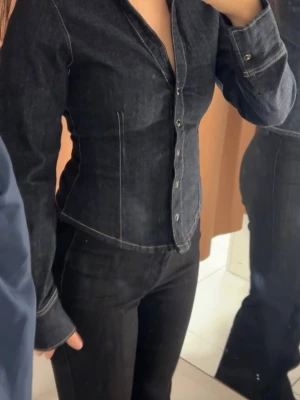 Mörk jeansskjorta med knappar - Snygg mörkblå jeansskjorta med markerad midja och tydliga sömmar. Skjortan har långa ärmar, v-ringning och knappar framtill. Perfekt för en trendig och edgy look. Helt ny med prislappen kvar!