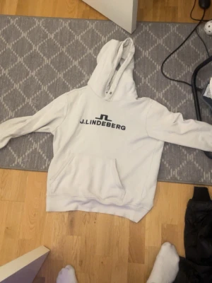 Vit hoodie från J.Lindeberg - Säljer en stilren vit hoodie från J.Lindeberg med svart logga och text på bröstet. Hoodien har en klassisk känguruficka framtill och en stor huva. Perfekt för dig som gillar enkel och clean streetwear även för dig som spelar golf en sen sommar kväll 🤩. Pris kan sänkas vid snabb affär