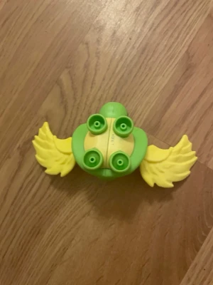 McDonald’s leksak ifrån adopt me collaboration  - Rolig plastleksak i grönt och gult med två stora vingar. Formen påminner om ett djur eller en fantasifigur och den har fyra fästen på undersidan. Perfekt till lek eller som del i ett byggset.