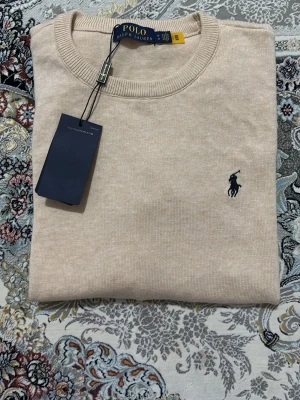 Beige stickad tröja från Polo Ralph Lauren - Snygg beige stickad tröja från Polo Ralph Lauren med klassisk rund halsringning och mörkblå broderad logga på bröstet. Tröjan har ribbade muddar och är tillverkad i mjukt material som passar perfekt till lager på lager.