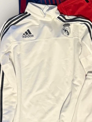 Vit Real Madrid träningströja Adidas - Snygg vit träningströja från Adidas med Real Madrid-logga på bröstet och svarta ränder längs ärmarna. Hög krage och dragkedja upptill. Perfekt för fotbollsträning eller chill. Klassisk sportig look med ikoniska detaljer.