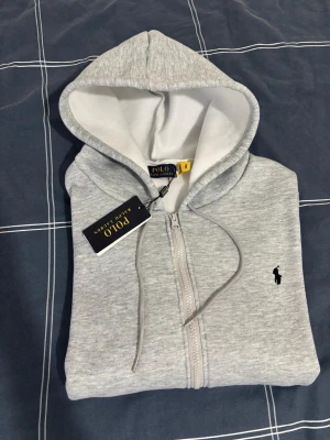 Grå zip hoodie från Polo Ralph Lauren - Snygg ljusgrå hoodie från Polo Ralph Lauren med dragkedja och klassisk svart logga broderad på bröstet. Hoodien har vit insida i huvan och justerbara snören. Perfekt för en chill och stilren look.