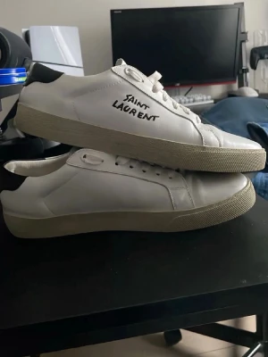 Saint Laurent vita sneakers - Snygga vita sneakers från Saint Laurent med svart detalj på hälen och handskriven logga på sidan. Klassisk låg modell med vit sula och ovandel i läder. Perfekta för dig som gillar stilrena och exklusiva skor.
