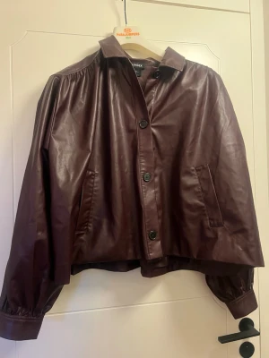 Vinröd skinnjacka från Lindex - Snygg vinröd skinnjacka från Lindex med oversized passform, knäppning framtill och två fickor. Jackan har ballongärmar med breda muddar och klassisk krage. Perfekt för dig som vill sticka ut med en trendig look.