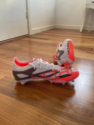 Puma Ultra fotbollsskor vit/röd - Säljer ett par Puma Ultra fotbollsskor med snygg design i vitt, rött och grått. Skorna har låg profil, lätta material och klassiska dobbar för bra grepp på planen. Perfekta för dig som vill ha snabbhet och stil på fotbollsplanen.
