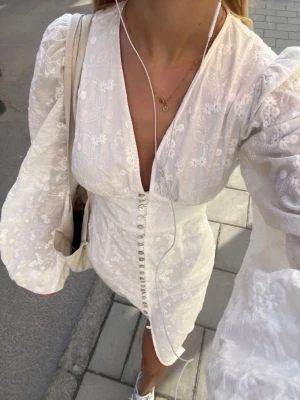 Vit långklänning med puffärmar - Superfin vit långklänning med v-ringning, puffiga långärmar och knappar framtill. Klänningen har broderade blommor i tyget och en slits framtill för extra stilpoäng. Klänningen har knappar framtill och en slits. En knapp fattas längst ner men märks knappt(Se första och 3e bild) Från adoore Strl 34 