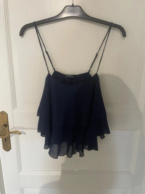 Mörkblå volangtop från Gina Tricot - Säljer en mörkblå volangtop från Gina Tricot med tunna justerbara axelband. Toppen har dubbla lager volanger. 