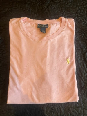 Rosa t-shirt från Polo Ralph Lauren - Säljer en stilren rosa t-shirt från Polo Ralph Lauren med klassisk gul broderad logga på bröstet. T-shirten har rund halsringning och är tillverkad i mjuk bomull, perfekt för en avslappnad och trendig look. Varan skickas ut dagen eller dagen efter köpet.