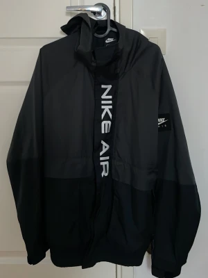 Svart Nike Air vindjacka med huva - Snygg svart vindjacka från Nike Air med stor logga framtill och huva. Jackan har dragkedja, elastiska muddar och en stilren design. Perfekt för blåsiga dagar och streetwear-stil. Lite trasig på framsidan men det syns inte så mycket.